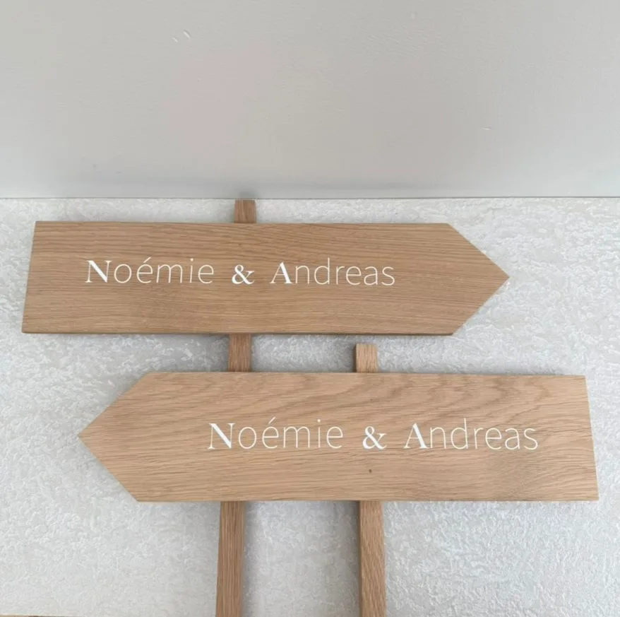 Twee houten wegwijzers in eik met de namen Noémie & Andreas in witte letters, signalisatie voor het huwelijk door Atelier V